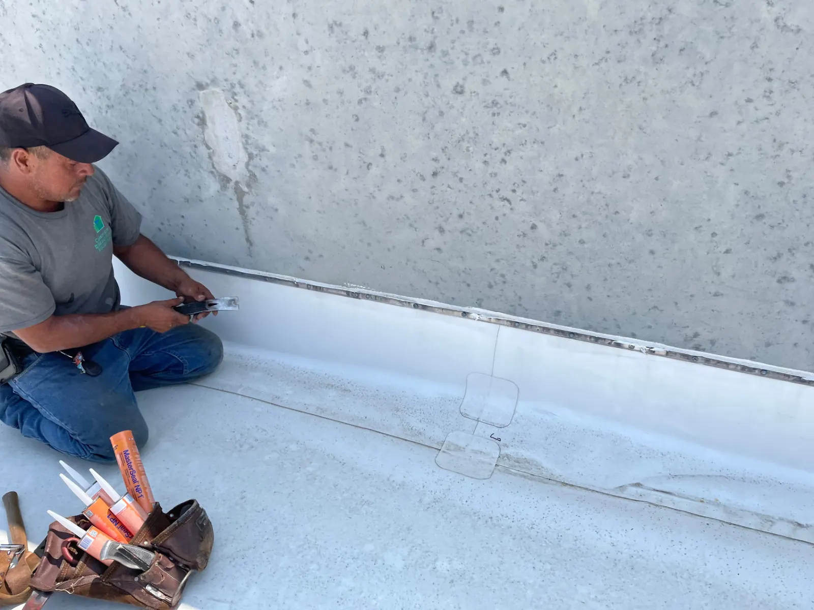 Commercial roofing crew member fitting a termination plate along a parapet base with MasterSeal NP1 sealant tubes in his tool bag