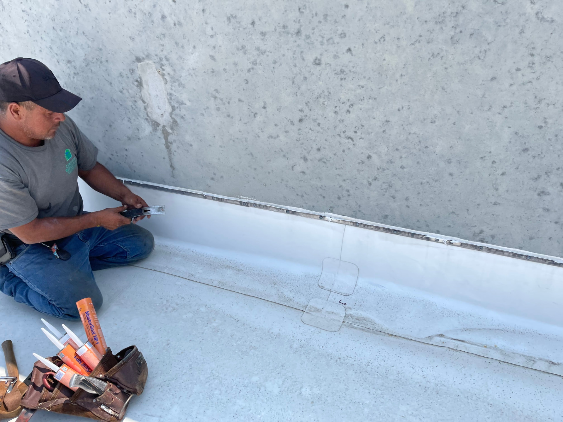 Commercial roofing crew member fitting a termination plate along a parapet base with MasterSeal NP1 sealant tubes in his tool bag